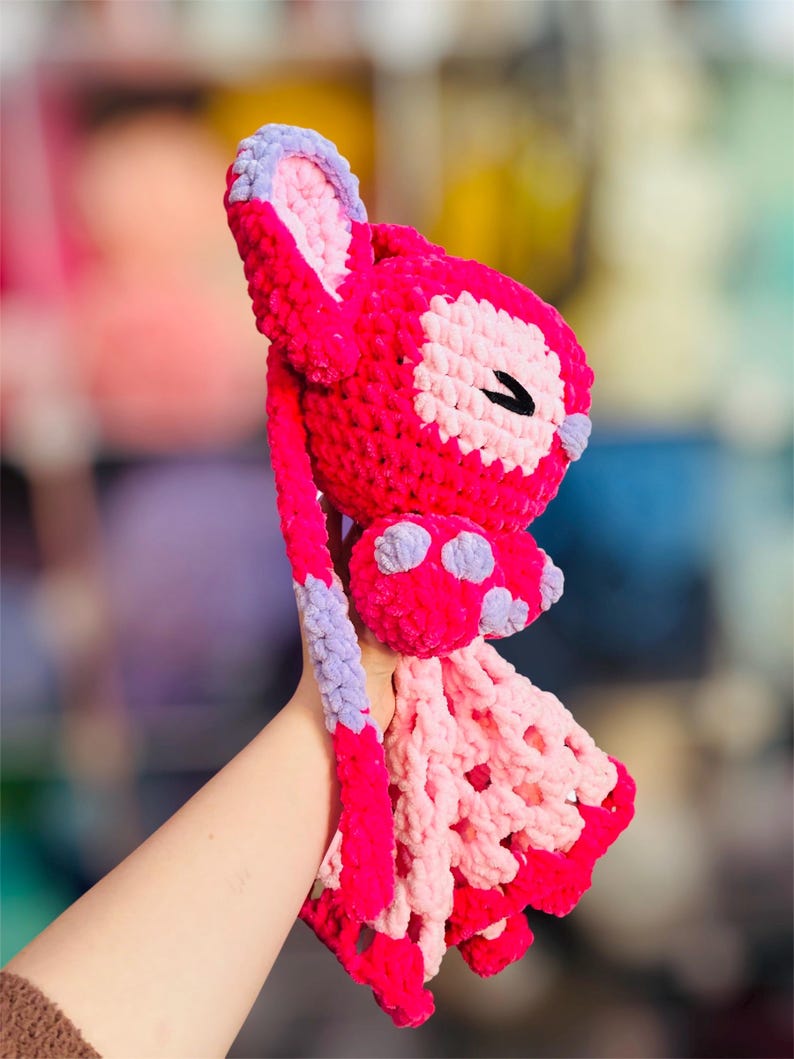 May include: A handmade, crochet bunny in shades of pink, lavender, and white. The bunny features long ears, a bow with purple polka dots, and a decorative openwork skirt. The bunny is held up by a person's hand.