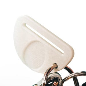 Op de afbeelding: Een witte, 3D-geprinte sleutelorganizer met een halfrond ontwerp en een rechthoekige sleuf. De organizer is bevestigd aan een zilveren sleutelring. De sleutelorganizer is ontworpen om sleutels te bewaren en te organiseren.