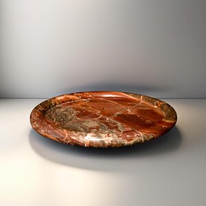 Può includere: Un vassoio decorativo rotondo con finitura lucida. Il vassoio presenta un colore marrone-rossastro con intricati motivi venati nei toni del bianco e del marrone. Il design suggerisce che sia fatto di pietra o materiale simile.