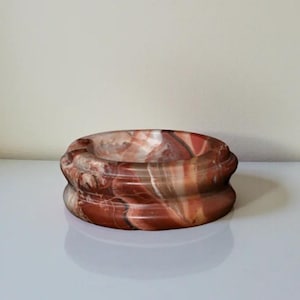 Può includere: Una ciotola decorativa rotonda in pietra rossa e crema. La ciotola ha un design a strati con una finitura liscia e lucida. I motivi naturali della pietra creano un aspetto marmorizzato unico. La ciotola è adatta per l'arredamento della casa.