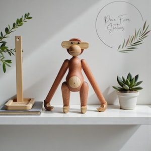 Op de afbeelding: Een houten apenbeeldje met beweegbare ledematen en een lichtbruine afwerking. De aap heeft een ronde kop, grote oren en een cirkelvormig buikdetail. De tekst "Pure Form Statues" staat in een cirkel op de achtergrond.