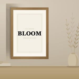 Op de afbeelding: Een rechthoekige wandlijst met een lichtbeige rand en een witte print. De print bevat het woord BLOOM in dikke zwarte hoofdletters, met kleinere tekst erboven en eronder: DAILY REMINDER en WHERE YOU ARE PLANTED.