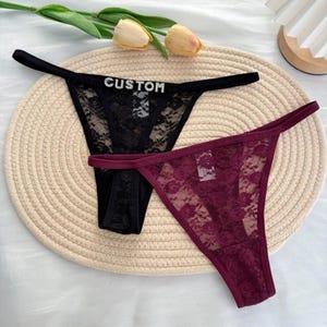 May include: Two thong underwear pieces. One is black with the word "CUSTOM" in rhinestones, and the other is burgundy. Both are made of floral lace.