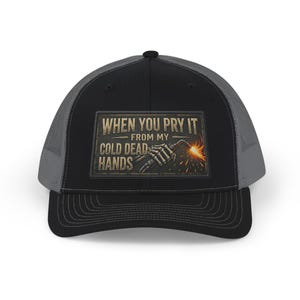 May include: A black and grey trucker cap featuring a rectangular patch on the front. The patch displays a skeleton hand holding a welding torch with sparks and the text: WHEN YOU PRY IT FROM MY COLD DEAD HANDS in a distressed, vintage-style font.