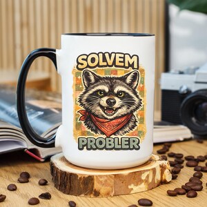 Könnte beinhalten: Weiße Keramiktasse mit schwarzem Henkel und Rand, mit einer Grafik eines Waschbären mit rotem Halstuch. Der Text auf der Tasse lautet "SOLVEM PROBLER" im Vintage-Stil. Die Tasse steht auf einem Holzuntersetzer, mit Kaffeebohnen verstreut.