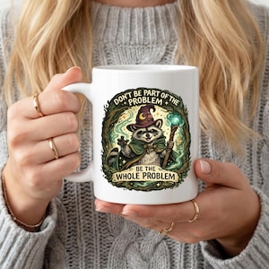 May include: White ceramic mug with a raccoon wizard design, holding a glowing staff. The mug features the text "DON'T BE PART OF THE PROBLEM BE THE WHOLE PROBLEM" within a decorative border. A quirky and amusing design.