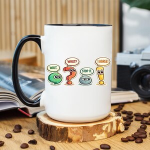May include: A white ceramic mug with a black handle and interior rim. The mug features a graphic design of four colourful punctuation marks with faces and speech bubbles containing the text: WAIT., WHAT?, STOP IT., and I AGREE!.