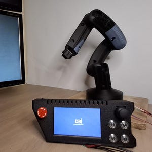 May include: A black robotic arm with a gray upper arm is positioned on a wooden surface. A control panel with a screen and red button is in the foreground. The screen displays the "DBi" logo. A computer monitor is in the background.