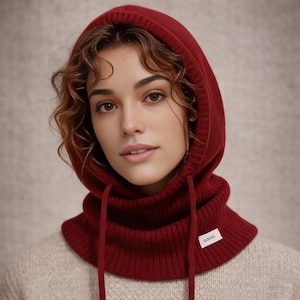 Puede incluir: Una capucha de punto color burdeos con cierre de cordón y cuello acanalado. La capucha se lleva sobre un suéter de color claro. Una pequeña etiqueta blanca con texto es visible en el cuello. La capucha está diseñada para mantener la cabeza y el cuello calientes.