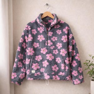 Pode incluir: Um pulôver de lã cinza com um padrão floral rosa. O pulôver tem um fecho de meio zíper, um bolso frontal e um cordão na bainha. A peça de roupa está pendurada num cabide de madeira.