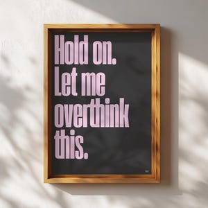 Può includere: Una stampa incorniciata con la scritta "Hold on. Let me overthink this." in rosa su sfondo nero. La cornice è in legno marrone chiaro. La stampa è esposta su una parete bianca.