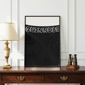 Puede incluir: Un estampado enmarcado con la palabra "Guinness" en blanco, con una fuente de burbujas sobre un fondo negro. La parte superior es blanca. El estampado se exhibe sobre una mesa de madera oscura.