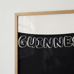 Puede incluir: Impresi&oacute;n enmarcada con la palabra "Guinness" en letras blancas redondeadas sobre un fondo negro. La parte superior es blanca y la impresi&oacute;n est&aacute; dentro de un marco de madera marr&oacute;n claro.