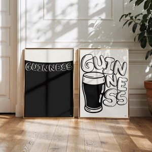 Puede incluir: Dos impresiones art&iacute;sticas enmarcadas con marcos dorados. Una presenta la palabra "Guinness" en blanco, sobre un fondo negro. La otra impresi&oacute;n muestra una pinta de Guinness con la palabra "Guinness" en una fuente de burbujas. Las impresiones se exhiben en un suelo de madera.