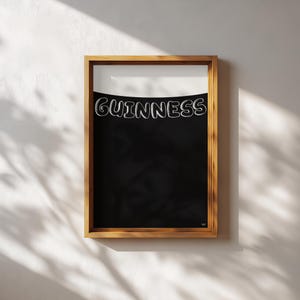 Puede incluir: Impresi&oacute;n enmarcada con el logotipo de Guinness en letras blancas curvadas sobre un fondo negro. La parte superior es blanca y el marco es de madera. La impresi&oacute;n est&aacute; colgada en una pared blanca, con sombras.