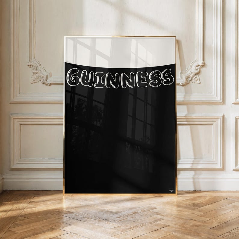 Puede incluir: Un cuadro enmarcado con la palabra "GUINNESS" en letras blancas estilizadas sobre un fondo en blanco y negro. La obra de arte tiene un marco dorado y se exhibe en una pared blanca.