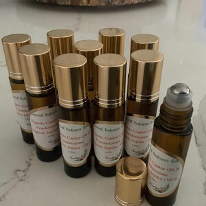 May include: A collection of amber glass roll-on bottles with gold caps. Each bottle has a label with text, including "Organic Castor Oil & Frankincense Jojoba". The bottles are arranged on a light-colored surface.