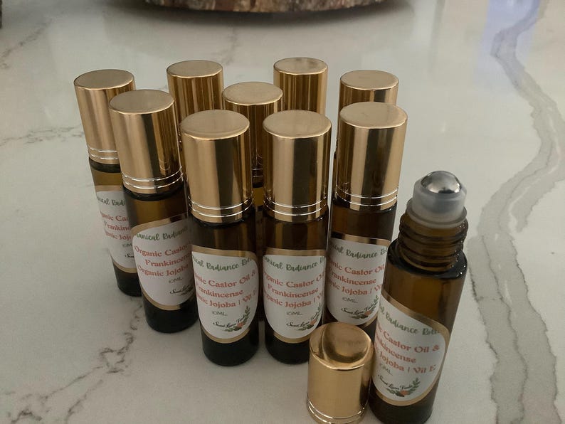 May include: A collection of amber glass roll-on bottles with gold caps. Each bottle has a label with text, including "Organic Castor Oil & Frankincense Jojoba". The bottles are arranged on a light-colored surface.