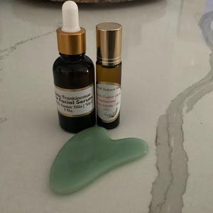 May include: A skincare set featuring a dark amber glass bottle with a dropper, a small roller bottle, and a jade green gua sha tool. The bottles have gold-colored caps and labels. The facial serum bottle is labeled "Frankincense Facial Serum".