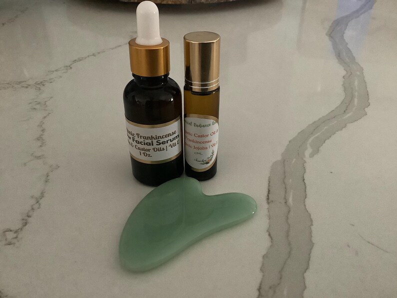 May include: A skincare set featuring a dark amber glass bottle with a dropper, a small roller bottle, and a jade green gua sha tool. The bottles have gold-colored caps and labels. The facial serum bottle is labeled "Frankincense Facial Serum".