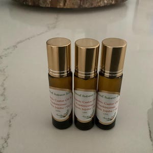 May include: Three amber glass roll-on bottles with gold caps. Each bottle has a white label with text that includes "Castor Oil, Frankincense, Jojoba". The bottles are arranged in a row on a light-colored surface.