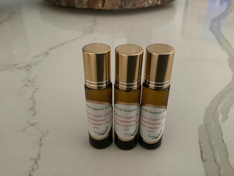 May include: Three amber glass roll-on bottles with gold caps. Each bottle has a white label with text that includes "Castor Oil, Frankincense, Jojoba". The bottles are arranged in a row on a light-colored surface.