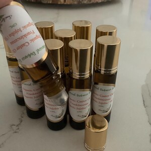 May include: A collection of small, brown glass roll-on bottles with gold-colored caps. Each bottle has a white label with text that includes "Organic Jojoba" and "Frankincense". The bottles are arranged on a light-colored surface.