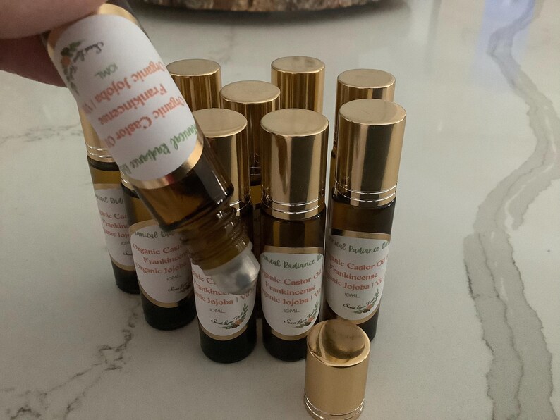 May include: A collection of small, brown glass roll-on bottles with gold-colored caps. Each bottle has a white label with text that includes "Organic Jojoba" and "Frankincense". The bottles are arranged on a light-colored surface.