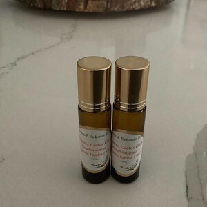 May include: Two small, amber-colored glass roll-on bottles with gold-colored caps. Each bottle has a label with text that includes "Essential Radiance Blend", "Castor Oil", "Frankincense", and "Jojoba". The bottles are on a white and gray marble-patterned surface.