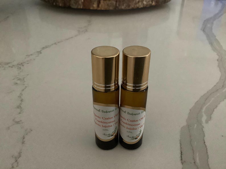 May include: Two small, amber-colored glass roll-on bottles with gold-colored caps. Each bottle has a label with text that includes "Essential Radiance Blend", "Castor Oil", "Frankincense", and "Jojoba". The bottles are on a white and gray marble-patterned surface.