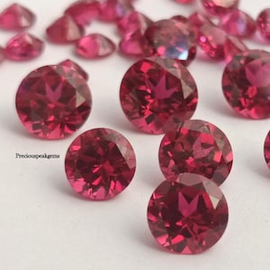 Op de afbeelding: Een verzameling gefacetteerde, ronde, dieprode edelstenen. De edelstenen zijn verspreid over een wit oppervlak, sommige op de voorgrond en andere op de achtergrond. De edelstenen hebben een briljante slijpvorm en reflecteren licht.