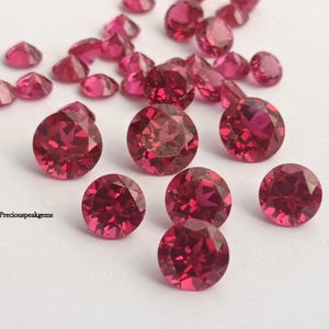 May include: A collection of faceted, round, deep red gemstones scattered on a white surface. The gems vary in size, with some appearing as small triangles and others as larger circles. The gems have a brilliant, sparkling appearance.