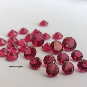 Może przedstawiać: Kolekcja fasetowanych, okrągłych i trójkątnych, głęboko czerwonych kamieni szlachetnych. Kamienie są rozrzucone na białej powierzchni, a światło odbija się od ich wypolerowanych powierzchni. Widoczny jest tekst "Preciouspeakgems".