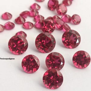 May include: A collection of faceted, round, deep pink gemstones scattered on a white surface. The gems vary in size, with some appearing more oval-shaped. The image includes the text "preciouspeakgems".