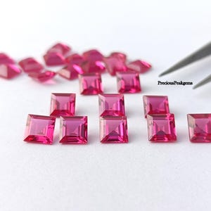 Può includere: Una collezione di gemme rosa vivaci, tagliate a forma quadrata. Le gemme sono disposte su una superficie bianca, alcune a fuoco e altre leggermente sfocate. Le pinzette sono visibili nell'angolo in alto a destra. Il testo "PreciousPeakgems" è visibile.
