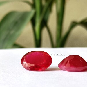 May include: Two faceted, oval-shaped, deep red gemstones. The gems are displayed on a white surface, with a blurred green plant in the background. The text "PreciousPeakGems" is visible on the right side of the image.