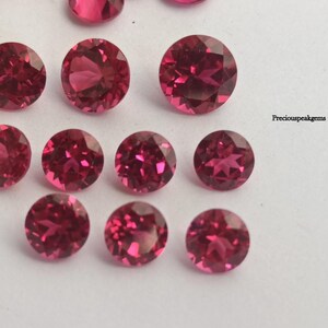 May include: A selection of faceted, round and oval-cut, deep pink gemstones. The gems are arranged on a white surface, showcasing their brilliant cut and colour. The image is well-lit, highlighting the sparkle and clarity of the stones. The text "preciouspeakgems" is visible.