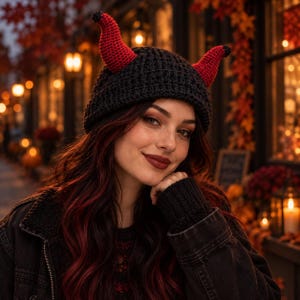 Può includere: Un berretto lavorato all'uncinetto di colore nero, caratterizzato da due corone rosse prominenti e dritte con punte nere. Il cappello è realizzato con un motivo a coste testurizzato ed è progettato per aderire perfettamente alla testa.
