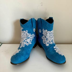 May include: A pair of bright blue ankle boots featuring white floral lace embroidery on the outer sides and toe areas. The boots have a rounded toe, a low block heel, and a pull-on style with side tabs. The material has a matte, textured finish.