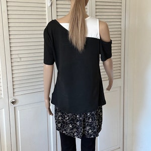 以下が含まれることがあります： 肩出しの黒と白の重ね着トップス。トップスは下に花柄のプリントスカートが付いています。モデルは黒いレギンスを履いています。
