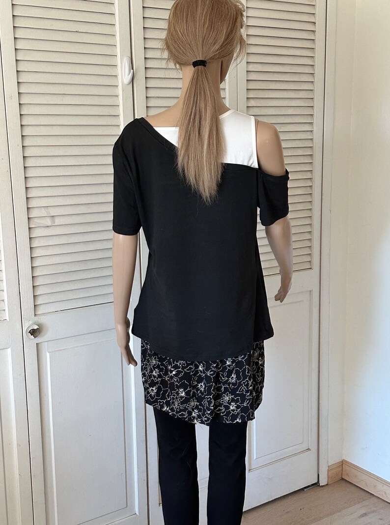 以下が含まれることがあります： 肩出しの黒と白の重ね着トップス。トップスは下に花柄のプリントスカートが付いています。モデルは黒いレギンスを履いています。