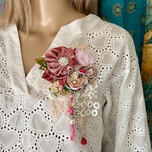 Peut inclure: Une broche florale avec une grande fleur en velours rose poudré et un centre en strass. La broche comprend de plus petites fleurs roses et crème, des perles et des chaînes pendantes. La broche est fixée à une chemise blanche à œillets.