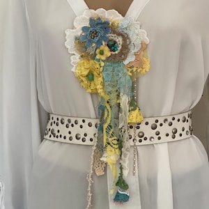 Peut inclure: Une broche florale multicolore avec des accents de dentelle et de tissu bleus, jaunes et crème. La broche présente un bouquet de fleurs et des rubans pendants. Une ceinture blanche cloutée est également visible.
