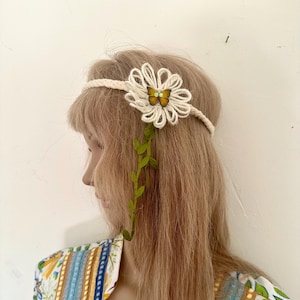 May include: A cream-colored headband with a large, crocheted flower and a small butterfly in the center. Green leaves cascade down the side. The headband is a bohemian style accessory.