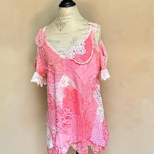 Puede incluir: Top de encaje rosa con diseño patchwork, expuesto en un maniquí. El top presenta una variedad de tonos rosas, aplicaciones florales y ribete de encaje blanco en las mangas y el escote. La prenda tiene un dobladillo asimétrico.