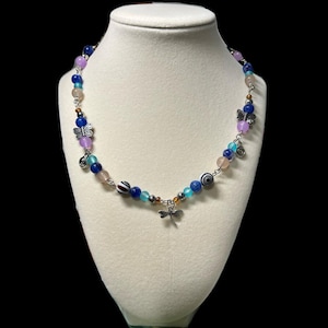 May include: A beaded necklace with a variety of colorful beads in shades of blue, pink, and purple. The necklace features silver-toned charms, including butterflies, spirals, and a dragonfly pendant. The necklace is displayed on a white mannequin bust.