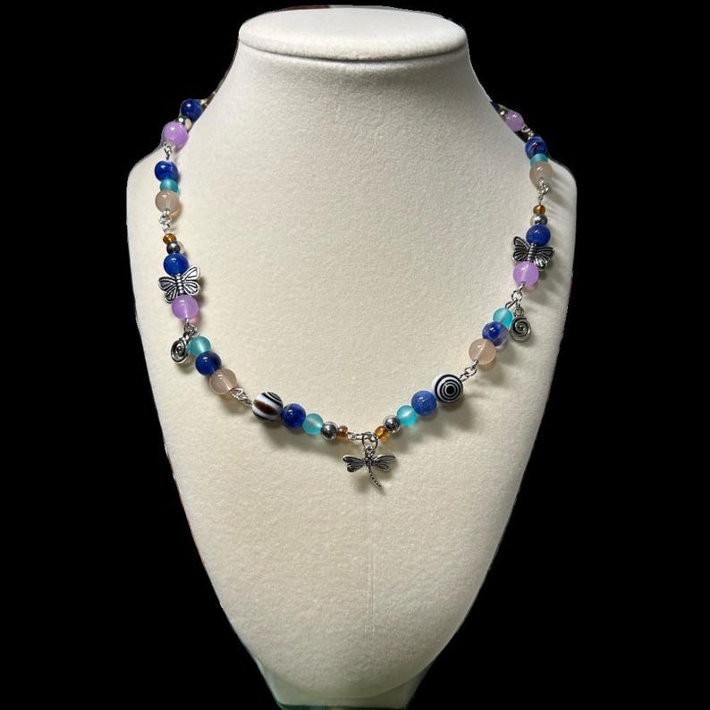 May include: A beaded necklace with a variety of colorful beads in shades of blue, pink, and purple. The necklace features silver-toned charms, including butterflies, spirals, and a dragonfly pendant. The necklace is displayed on a white mannequin bust.