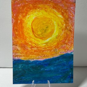 May include: A rectangular painting on a clear plastic stand featuring a vibrant yellow sun centered in a textured orange and pink sky above a deep blue sea. The artwork displays visible brushstrokes and layered colors, creating a bold, expressive landscape scene.