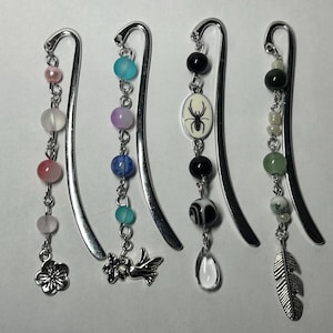 May include: Four metal bookmarks with decorative beaded charms. Charms include a flower, a bird, a spider, and a feather. The beads are in various colors, including pink, blue, green, and black. The bookmarks are on a white surface.