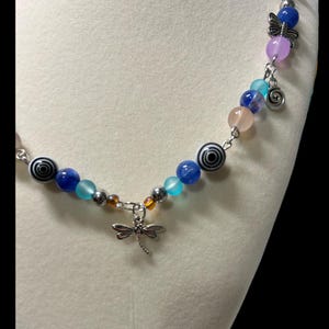 May include: A beaded necklace with a dragonfly pendant. The necklace features a variety of colorful beads, including blue, teal, pink, and black and white spiral beads. A silver dragonfly charm hangs from the center.